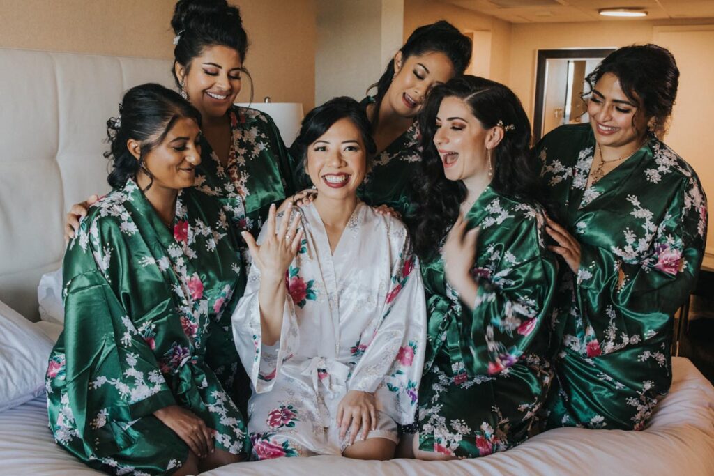 Bride sitting on a bed surrounded by her bridesmaids, all wearing matching green floral robes, as they celebrate and admire her engagement ring