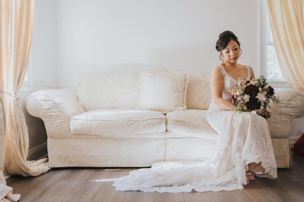 Bride sitting on a couch, holding her bouquet and looking thoughtfully down, with her wedding dress cascading on the floor at The Estate at Farrington Lake, East Brunswick, NJ
