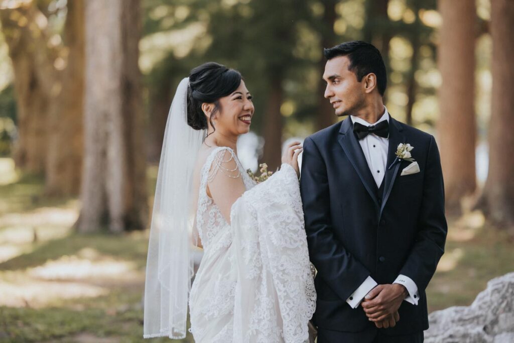 Bride playfully looking at the groom while holding her veil, with the groom smiling at her in a wooded area at The Estate at Farrington Lake, East Brunswick, NJ