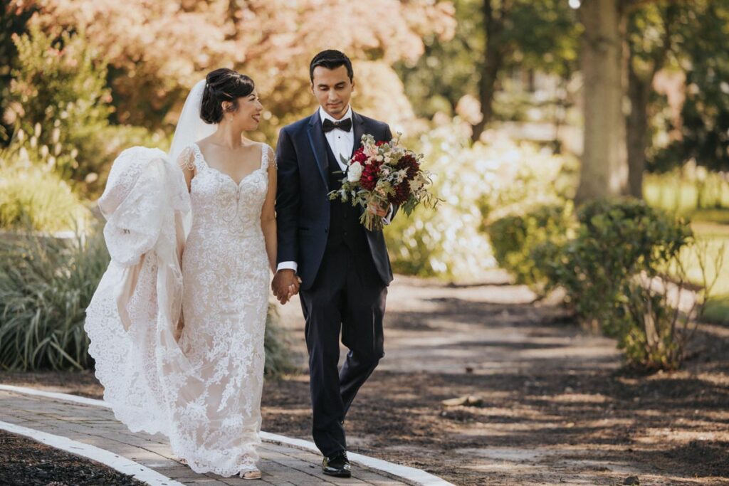 Bride and groom walking hand in hand along a path, smiling at each other, with the bride holding her bouquet at The Estate at Farrington Lake, East Brunswick, NJ