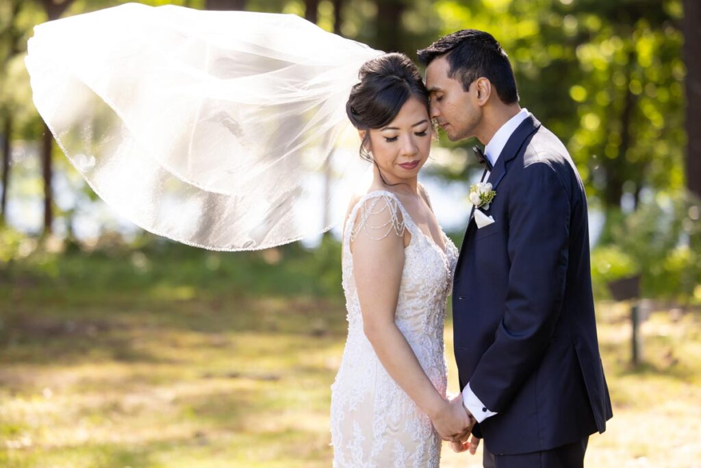 Groom resting his forehead against the bride's head as her veil flows in the wind at The Estate at Farrington Lake, East Brunswick, NJ