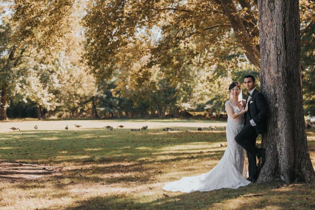 Bride and groom standing by a large tree, smiling in a sunlit field with geese in the background at The Estate at Farrington Lake, East Brunswick, NJ