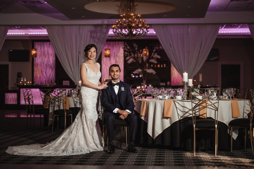 Bride standing beside the groom, who is seated, in an elegantly decorated reception area at The Estate at Farrington Lake, East Brunswick, NJ