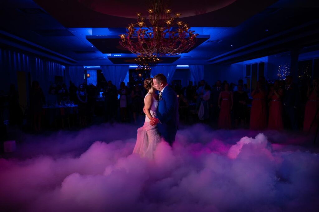 A couple shares a romantic dance on a cloud of mist, illuminated by colorful lights and a chandelier at The Estate at Farrington Lake, East Brunswick, NJ, surrounded by guests enjoying the celebration.