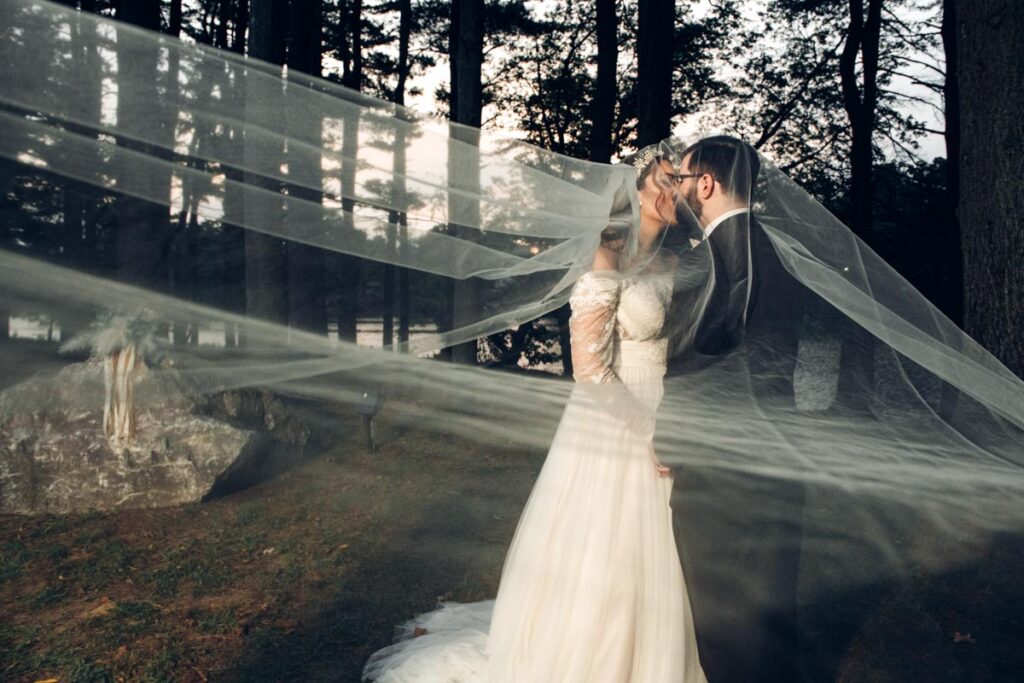Bride and groom kissing outdoors under a long flowing veil, surrounded by trees at dusk