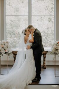 Bride and groom sharing a kiss during their wedding ceremony in front of a large window with floral arrangements