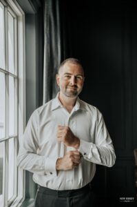 Groom standing by a window, adjusting his cufflinks and smiling