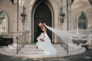 Bride and groom kissing on church steps, with the bride's veil flowing in the wind