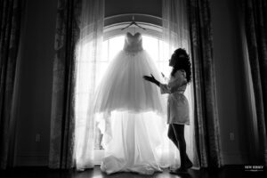 photography northern jersey - Bride in a robe admiring her wedding dress hanging in front of a large window