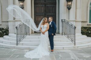 Bride and groom standing on church steps, with the bride's veil flowing in the wind