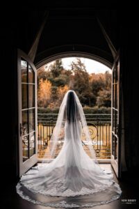 Bride standing in front of open doors, looking out onto a scenic outdoor view with her long veil trailing behind her