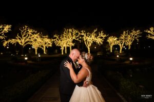 Bride and groom embracing at night with trees illuminated by string lights in the background