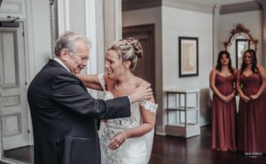 Bride sharing a joyful moment with her father, while two bridesmaids look on in the background
