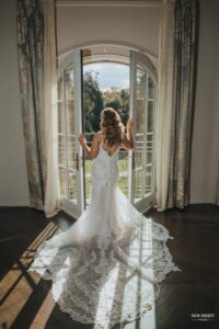 Bride standing in front of open doors, looking out, with her long lace train spread across the floor