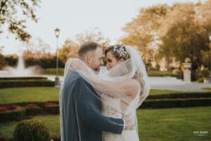 Bride and groom embracing outdoors, wrapped in the bride's veil, with a garden and fountain in the background