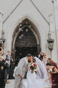 Bride and groom sharing a kiss outside a church as guests blow bubbles to celebrate