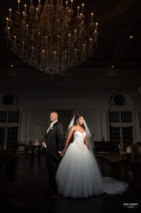Bride and groom standing back to back under a grand chandelier in a dimly lit ballroom