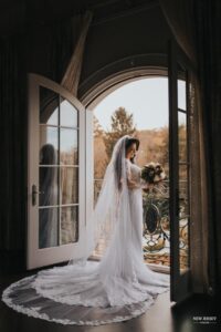 Bride standing by open doors, holding her bouquet, with her long veil and train trailing behind her