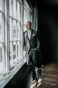 Groom in a suit leaning against a window, looking out thoughtfully