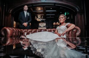 Bride sitting on a leather sofa with her dress elegantly spread out, while the groom stands behind her in a formal setting
