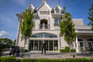 Groom standing outside in front of a large estate, while the bride looks out from a balcony above