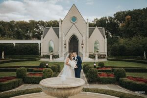 Bride and groom sharing a kiss in front of a small chapel, with a fountain and manicured gardens in the foreground
