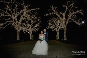 Bride and groom posing together at night, with trees wrapped in string lights in the background