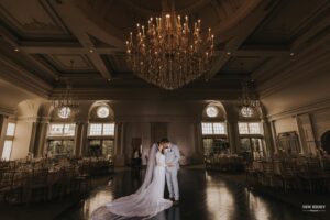 Bride and groom sharing a moment in an empty ballroom under a large chandelier, with the bride’s veil trailing on the floor