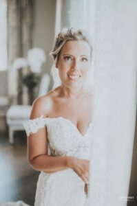 Close-up of a bride smiling softly while standing by a window in her wedding dress