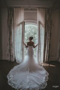 Bride standing in front of open doors, holding the curtains, with her lace train spread across the floor