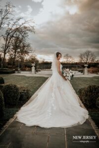 Bride standing outdoors, holding her bouquet, with her long train and veil flowing behind her